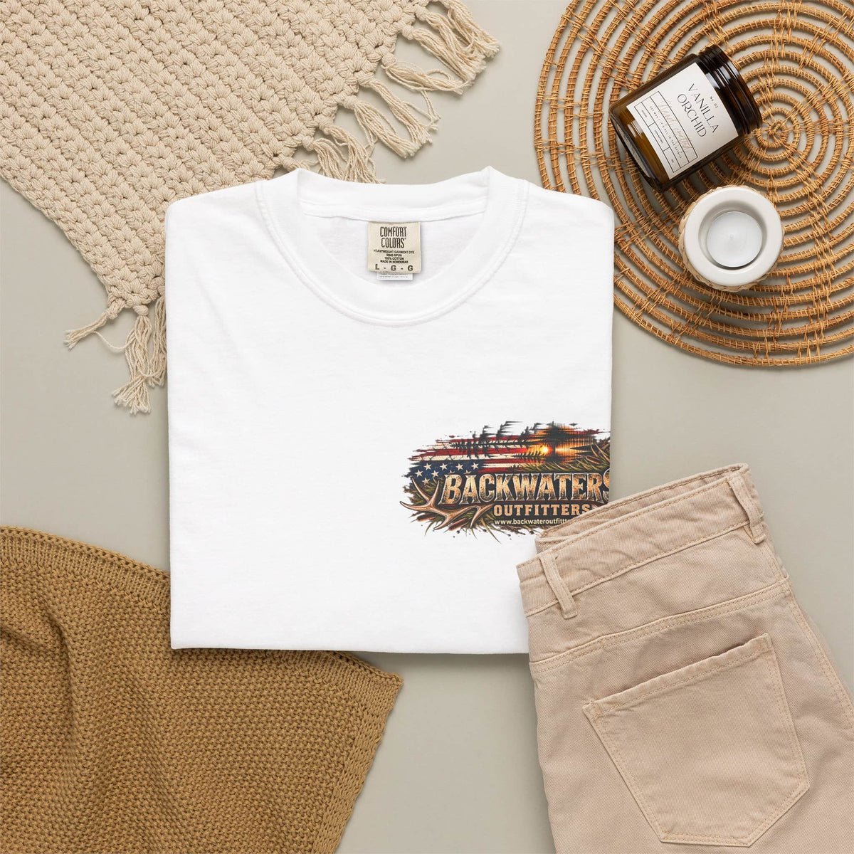 A folded white T-shirt is displayed in a styled flat lay with beige pants, a woven blanket, a candle jar, and a small cup on a neutral surface. The shirt has a front graphic reading Backwater Outfitters with a rustic American flag and sunset design.