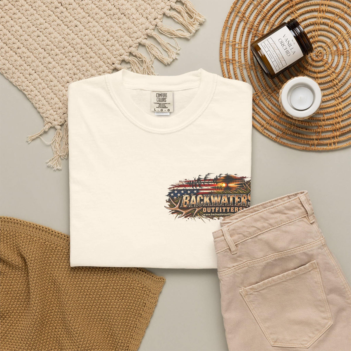 A folded off-white T-shirt is styled in a flat lay with beige pants, a woven blanket, a candle jar, and a small cup on a neutral surface. The shirt has a distressed graphic on the chest featuring an American flag and the text 'BACKWATER OUTFITTERS'.