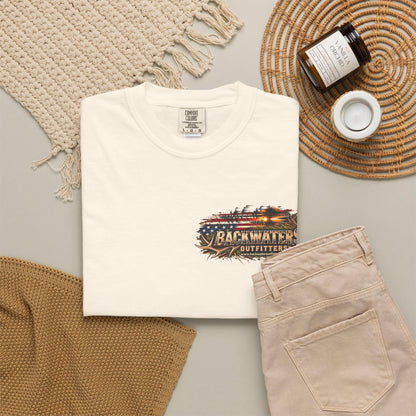 A folded off-white T-shirt is styled in a flat lay with beige pants, a woven blanket, a candle jar, and a small cup on a neutral surface. The shirt has a distressed graphic on the chest featuring an American flag and the text 'BACKWATER OUTFITTERS'.