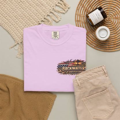 A folded light pink t-shirt with a 'Backwater Outfitters' graphic on the chest, styled in a flat lay with beige pants, a woven rug, a candle, and a jar on a neutral background.