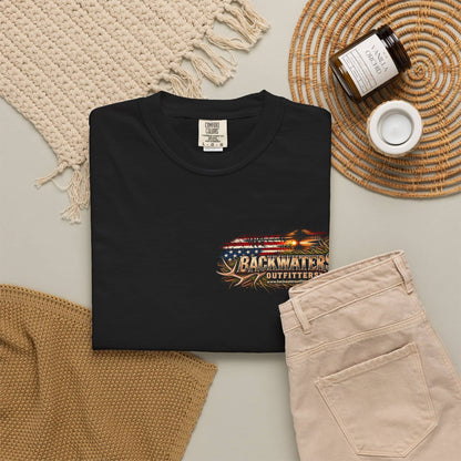 A folded black T-shirt is displayed in a styled flat lay with beige shorts, a woven rug, a candle jar, and a small cup on a neutral surface. The shirt has a colorful chest graphic reading 'BACKWATER OUTFITTERS' with an American flag and antler motif.
