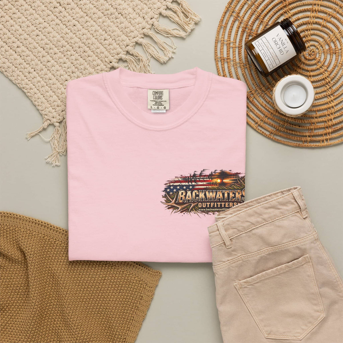 A folded light pink T-shirt is displayed in a styled flat lay with beige pants, a woven blanket, a candle jar, and a small cup on a neutral surface. The shirt has a front graphic reading Backwater Outfitters with an American flag and rustic wood design.