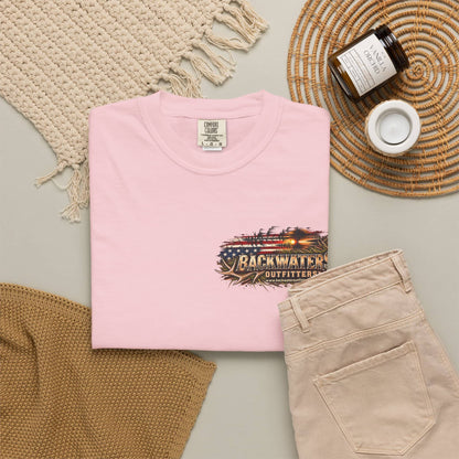A folded light pink T-shirt is displayed in a styled flat lay with beige pants, a woven blanket, a candle jar, and a small cup on a neutral surface. The shirt has a front graphic reading Backwater Outfitters with an American flag and rustic wood design.
