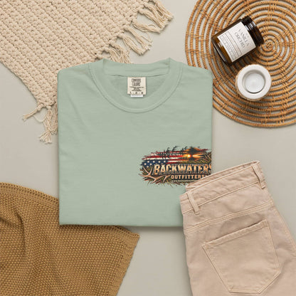 A folded sage green t-shirt with a rustic 'Backwater Outfitters' graphic on the chest, styled in a flat lay with beige pants, a woven blanket, a candle jar, and a small cup on a neutral background.