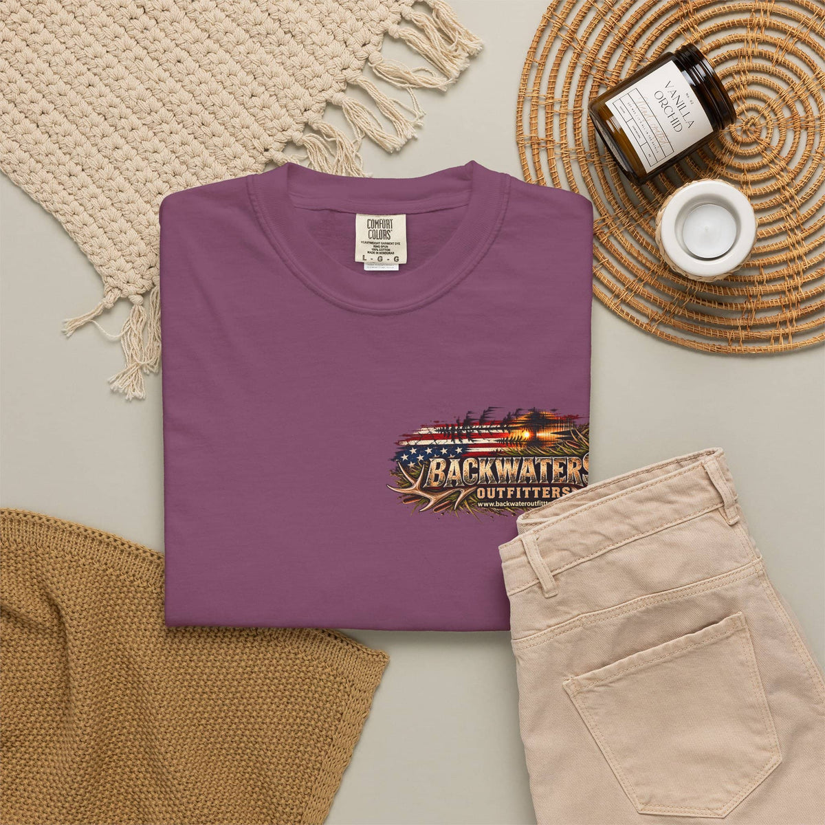 A folded purple crewneck t-shirt with a 'MOKI ISLAND' graphic on the left chest, styled in a flat lay with beige pants, a woven blanket, a candle, and a jar on a neutral background.