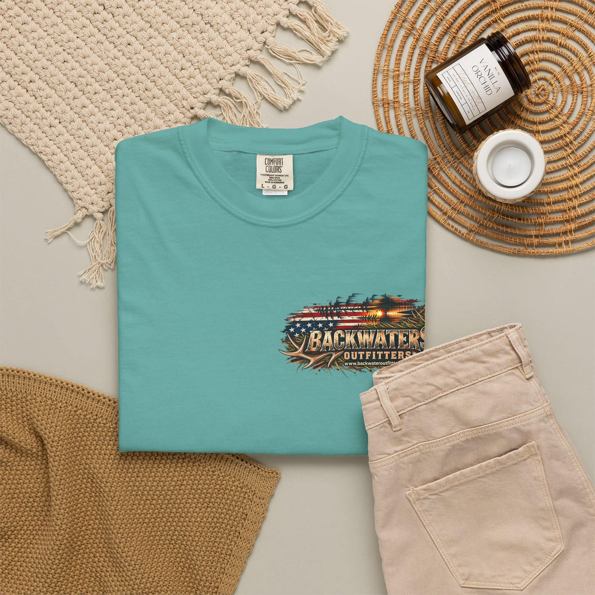 A folded turquoise crewneck t-shirt with a 'Backwater Outfitters' graphic on the chest, styled in a flat lay with beige pants, a woven blanket, a candle jar, and a small cup on a neutral background.