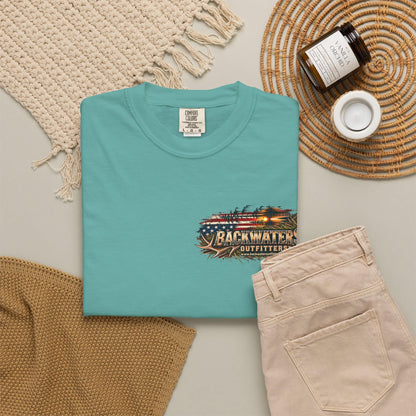 A folded turquoise crewneck t-shirt with a 'Backwater Outfitters' graphic on the chest, styled in a flat lay with beige pants, a woven blanket, a candle jar, and a small cup on a neutral background.